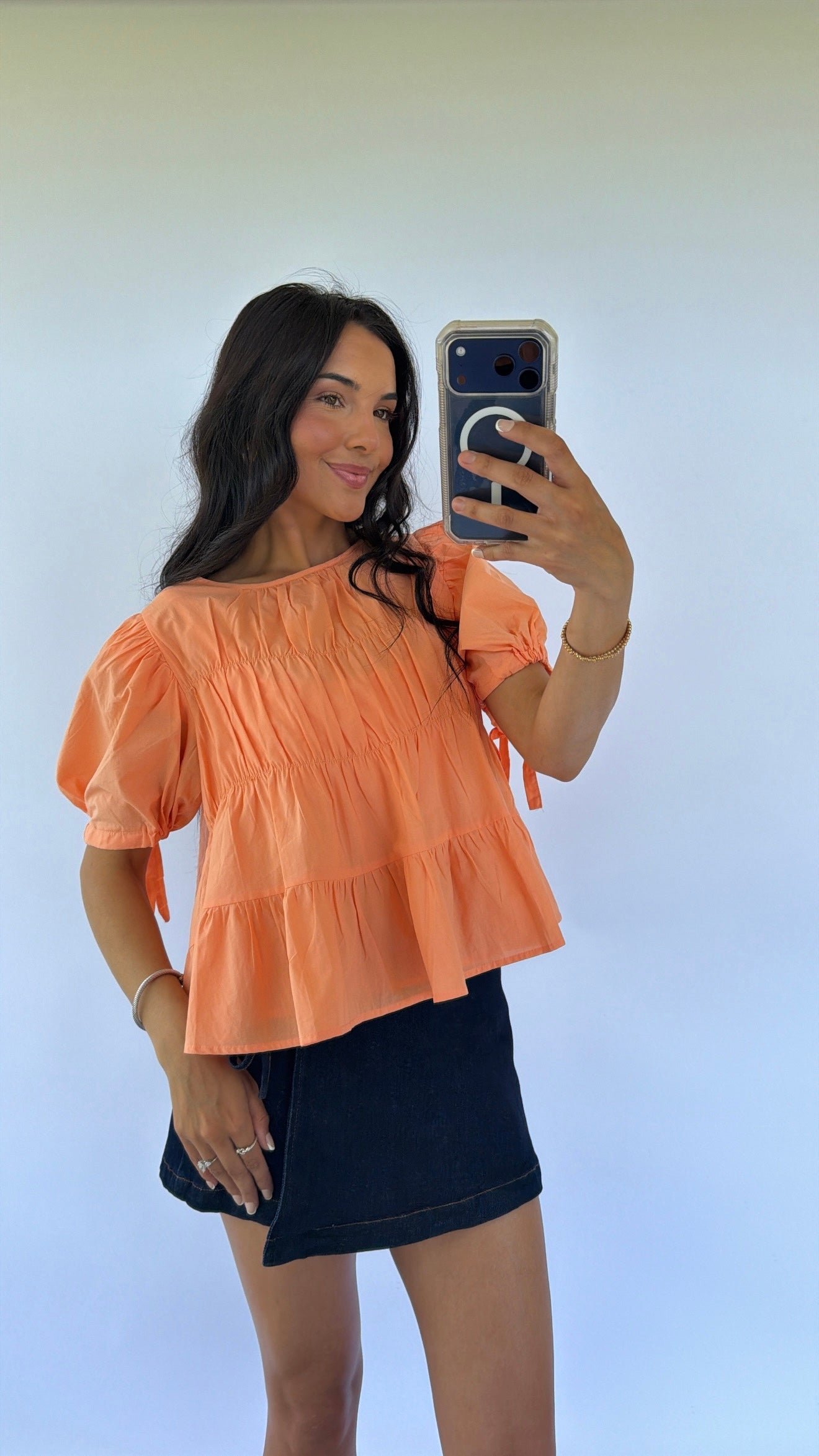 Woman taking a mirror selfie wearing an orange top and black skirt against a white background